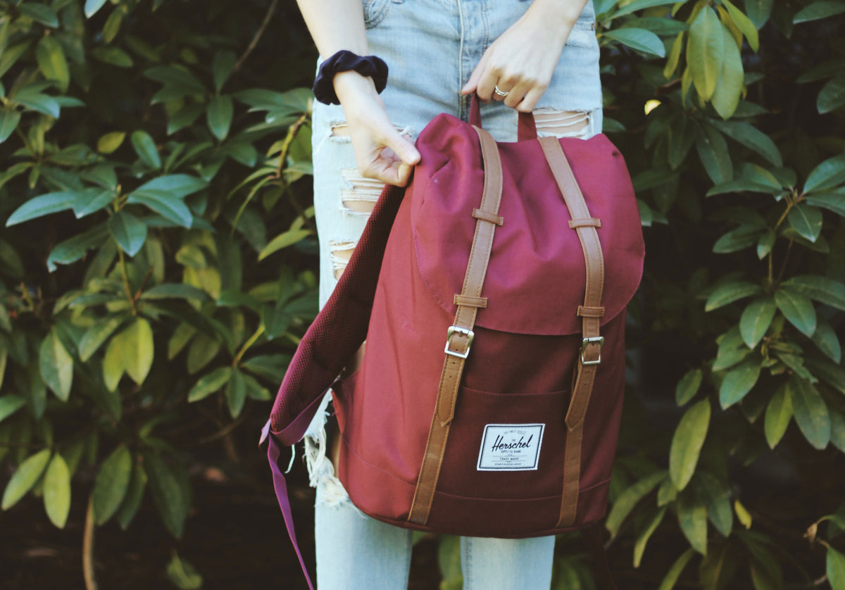 Person holding a burgundy Herschel backpack in front of green bushes.