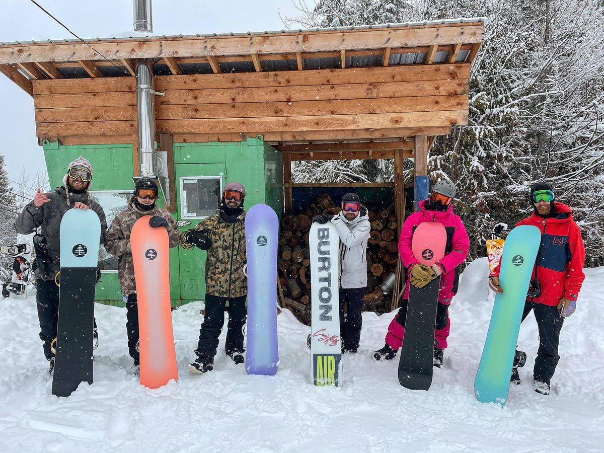 Group of snowboarders posing with colorful snowboards in front of a wooden mountain cabin.